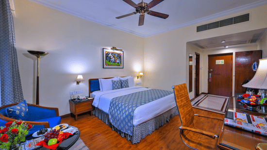 A room with a large bed, spreads, pillows, a fan, and windows at The Clarks Varanasi.
