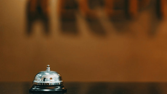 An image of a hand bell placed on a marble counter, with blurred, wall-mounted acrylic letters in the background.