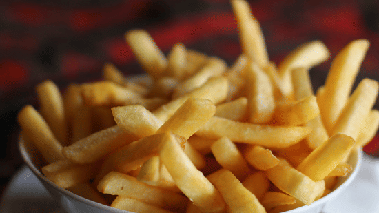 White bowl filled with golden, crispy French fries, freshly cooked with a glossy texture, set against a blurred dark red and black background.