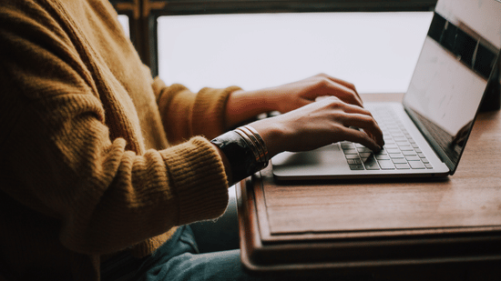 Side view of a person typing on a laptop computer set on a wooden table.