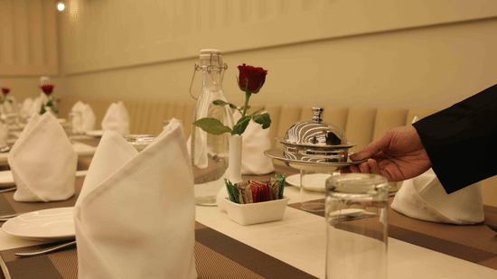 A close-up of a dining table at Tulip Inn Gurugram set with white napkins, a red rose in a vase, and a staff member's hand visible.