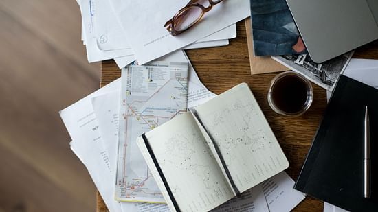 A cluttered wooden desk with papers, a notebook, glasses, a laptop, a drink, and other miscellaneous items