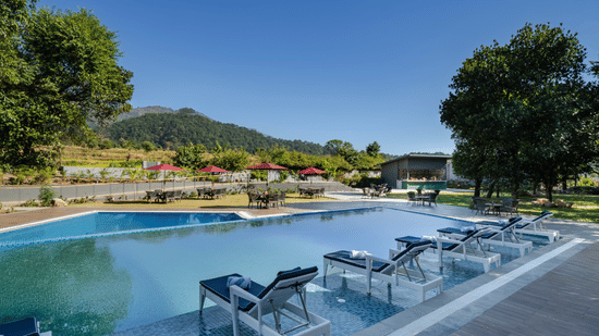 An outdoor swimming pool at Vandhara Sarovar Premiere, Patkote, with lounge chairs and mountain views under a clear sky.