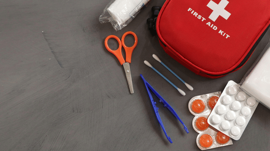 A first aid kit next to medical supplies including scissors, tweezers, cotton swabs, a bandage roll, and blister packs.