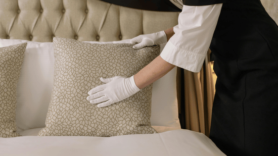 A person wearing white gloves adjusting a square patterned pillow on a bed with a tufted headboard.