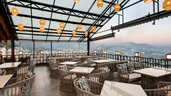An outdoor rooftop restaurant with a glass and metal roof, woven seating and tables, spherical pendant lights, and a panoramic view of the landscape | Voyage Eastin Suites