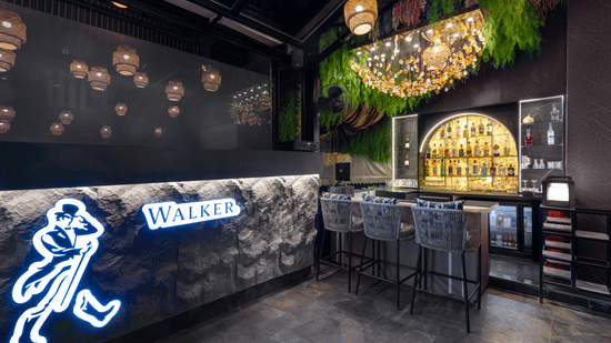 A dark, modern bar area with a lit counter, bar stools, bottles on shelving, and a neon sign of a walking character | Voyage Eastin Suites