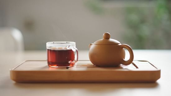 an image of a glass tea cup kept next to an earthen kettle on a tray inside a room