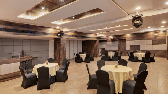 Spectrum hall at Voyage Milestone Hotel And Banquet featuring chairs arranged around round tables with flower vases on them, placed in distance under warm lighting and light equipment attached to the ceiling