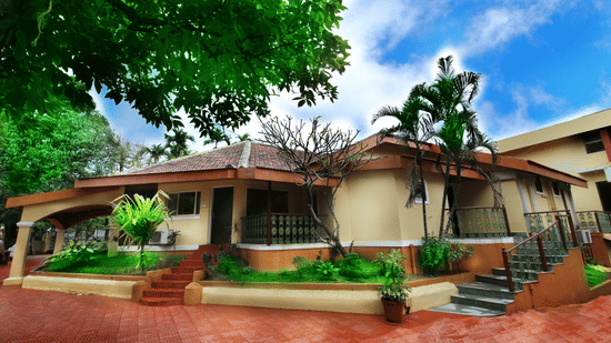 A single-storey cottage sits amid lush greenery, with tiled roofs, small stairways, and plants framing a quiet courtyard setting | Zara's Resort