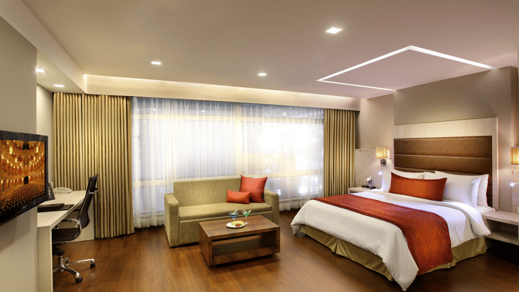 Premiere Suite at Mahagun Sarovar Portico Vaishali featuring a double size bed, a flat TV screen, a sofa with centre table, and a large window covered with curtains.