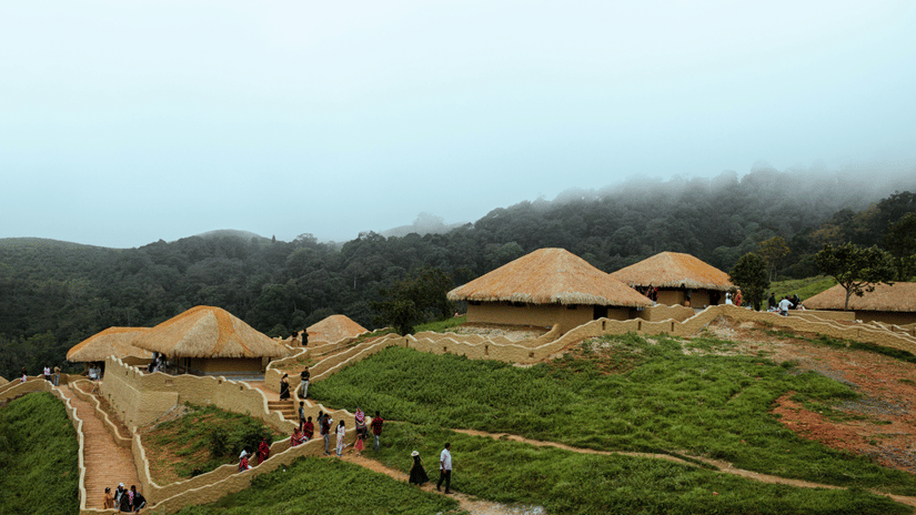 Earthen huts with thatched roofs built into a grassy hillside in En Ooru tribal village Wayanad, Kerala, with people walking on paths and mist covering the forest behind.