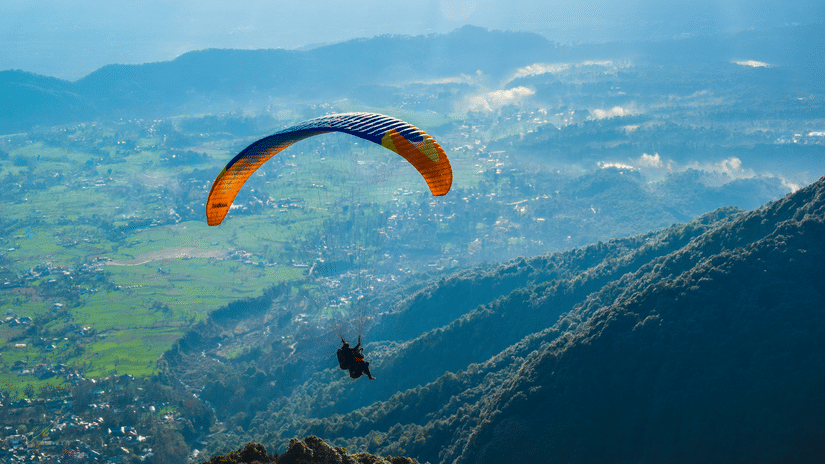 A paraglider with colourful canopy soaring above dramatic mountain ridges, with expansive green valleys and blue skies stretching beyond below