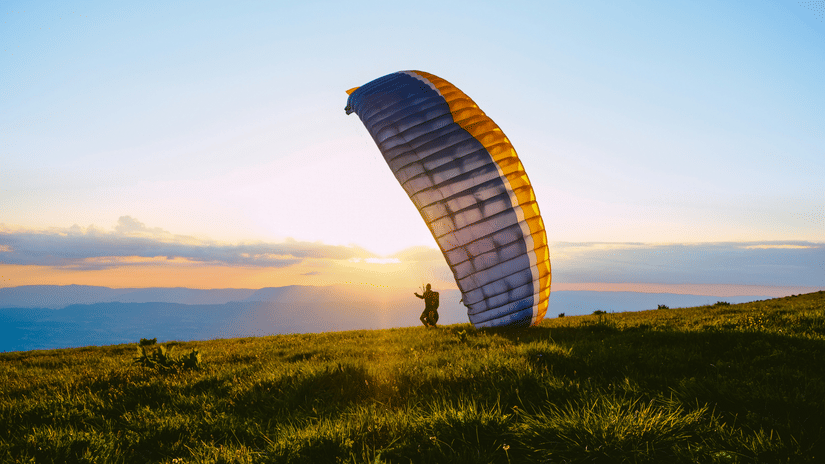 Colourful paraglider preparing for take-off across grassy hills, silhouetted against a warm sunrise with expansive open countryside beyond