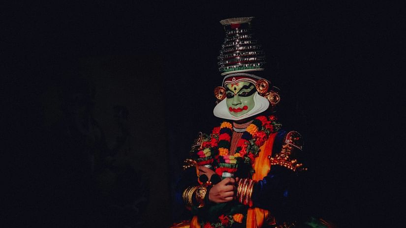A Kathakali dancer in elaborate costume and makeup poses against a dark background during a classical performance.