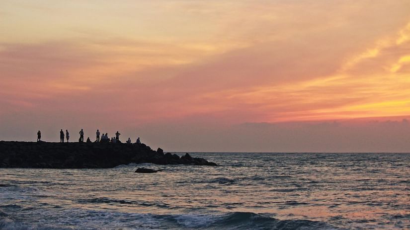 A vibrant orange and pink sunset over the ocean, with a silhouetted breakwater and figures in the distance.