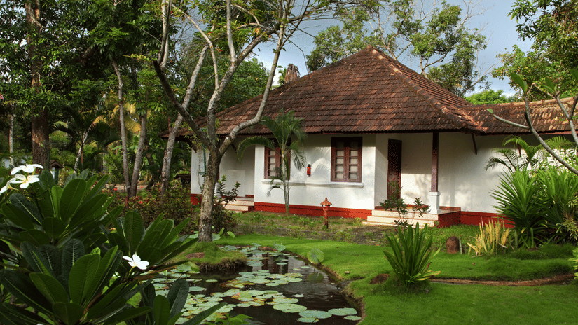 A traditional white cottage with a terracotta tile roof, red window frames, and a small porch, set in a lush tropical garden at Abad Turtle Beach Resort, Marari, Alleppey, an Alappuzha Beach Resort