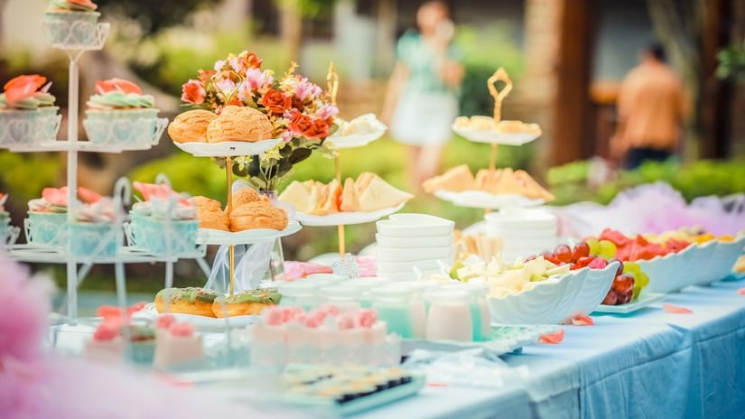 A blurred, outdoor view of a long table filled with a variety of snacks, pastries, fruit, and tiered displays of small desserts.