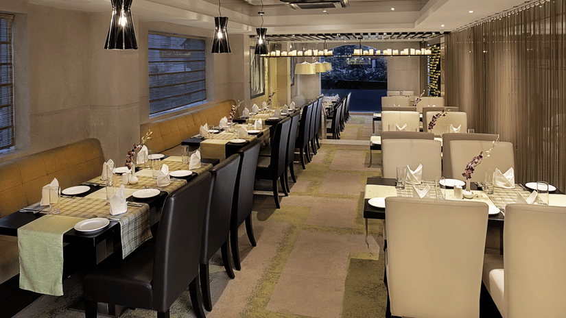 Interior view of Bahula restaurant at Barsana Boutique Hotel, Kolkata, showing organised rows of dining tables with neat place settings and contemporary decorative lighting.