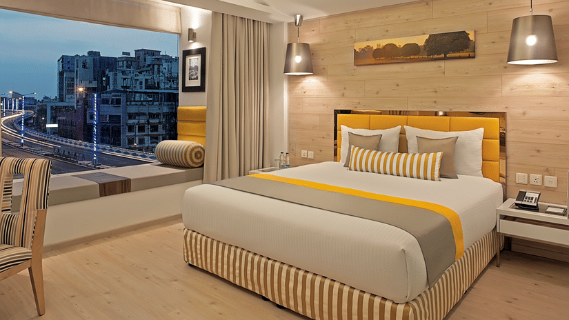 An elegantly designed Deluxe Room at Barsana Boutique Hotel, Kolkata, featuring a well-made bed along with a floor-to-ceiling window offering city views.