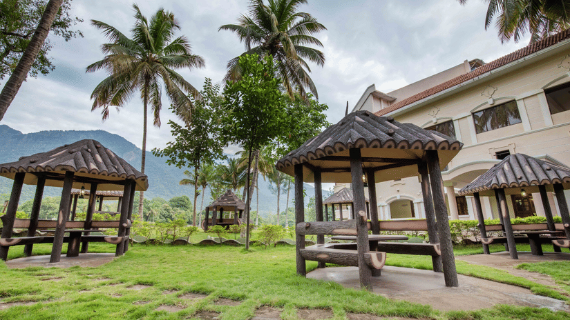 Grassy outdoor area with shaded wooden gazebos, ideal for relaxing, set among coconut trees with a view of the nearby resort - Black Thunder, Coimbatore
