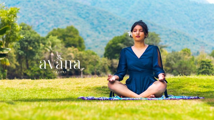 Woman practicing meditation on a yoga mat in a lush green field with scenic mountain views.