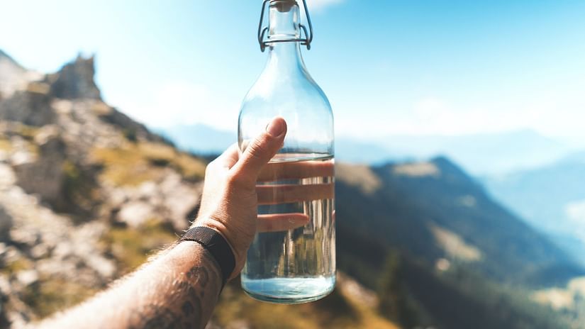 A person holding a reusable glass water bottle amidst mountains.