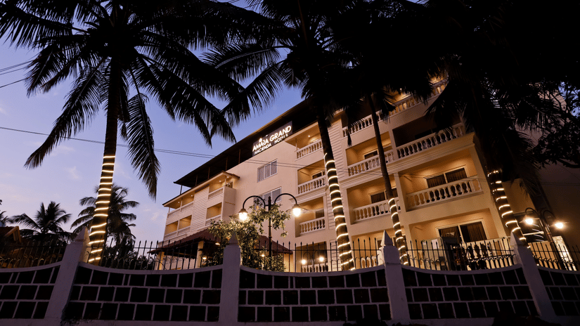 Exterior view of Amara Grand Baga, Goa, with a multi-storey building and trees on the side under a blue sky.