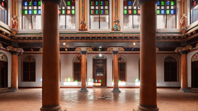 Long corridor with red and yellow tiled floor flanked by green potted plants and wooden columns at Chidambara Vilas Resort - The Best Resort in Chettinad.