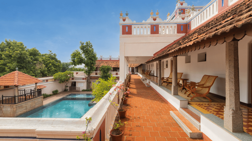 An outdoor view of Chidambara Vilas, a luxury heritage resort in Chettinad, featuring a long covered walkway with a terracotta floor, traditional architecture with accents, and an outdoor swimming pool surrounded by greenery under a  sky