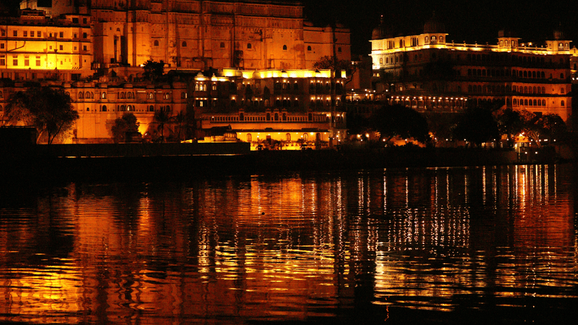 The night view of the banks of Ambrai Ghat in Udaipur, with the lake's water mirroring the beautiful wamly lit facade of the heritage buildings.