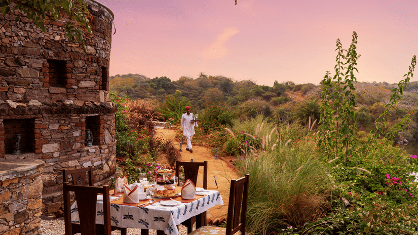 A table for 4 set up next to a stone building with forest cover in the backdrop at Chunda Shikar Oudi, Udaipur