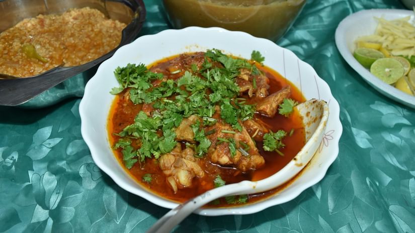 A rich, orange-red meat or chicken curry served in a white bowl, garnished heavily with fresh coriander, with other side dishes blurred in the background.