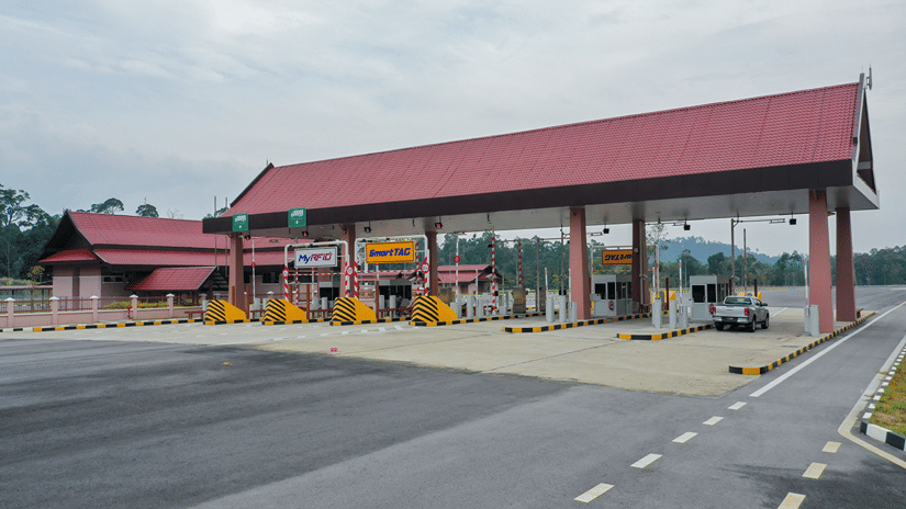 A red-roofed toll plaza with electronic payment lanes sits beside a quiet road under an overcast sky.