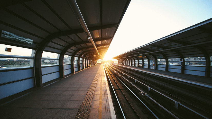 Bright sunlight glows at the end of an empty railway platform flanked by covered tracks beneath a clear sky.
