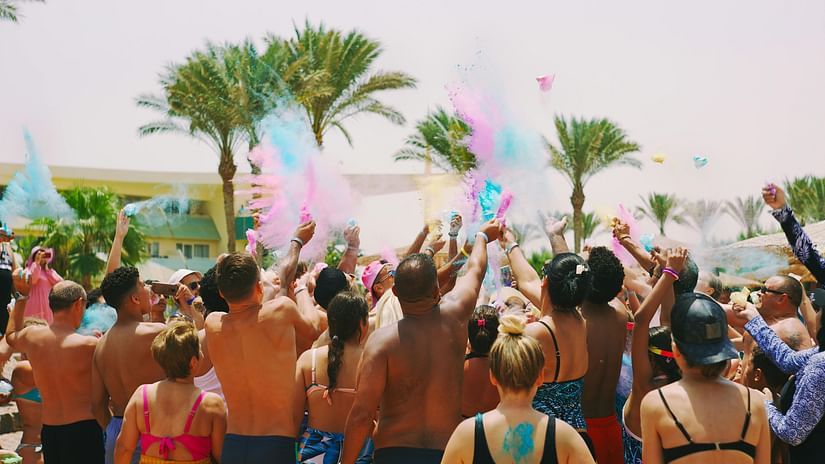 A crowd celebrates at a beach party, tossing vibrant Holi powder into the air.
