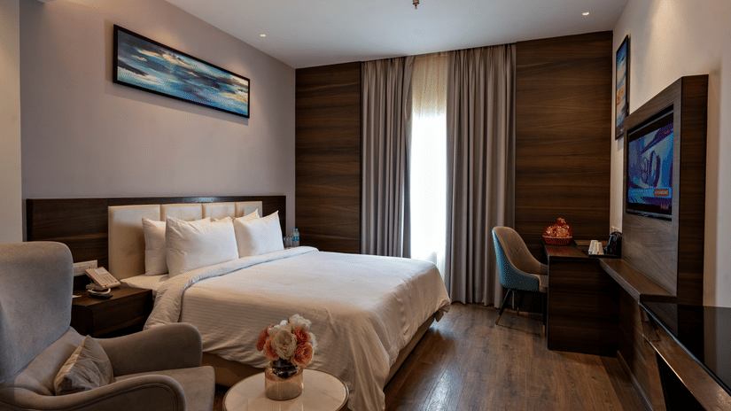 A hotel room at Geeta Sarovar Portico, Panipat featuring a bed, a flat TV screen mounted on the wall, a  chair and working desk, and a sitting area with arm chair and table.