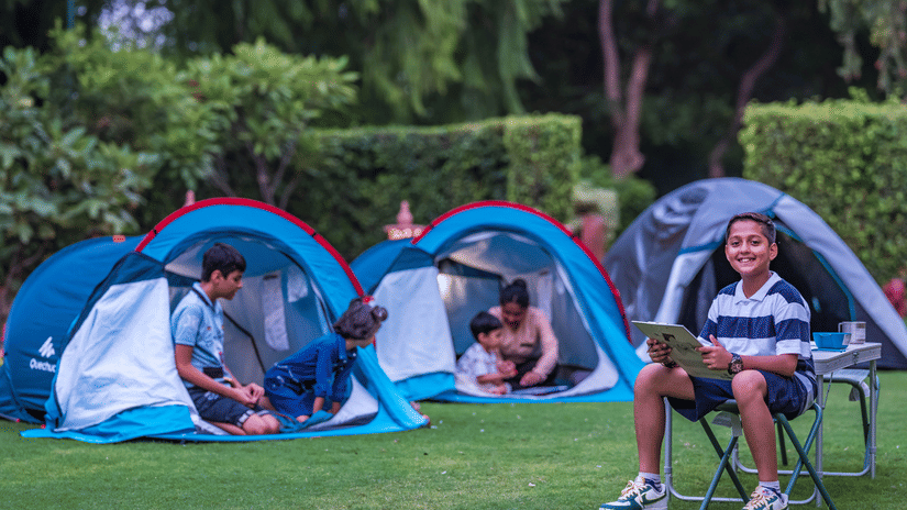 Children and adults sitting near blue tents on the lawn, involved in a camping activity at Heritage Village Resorts & Spa, Manesar.