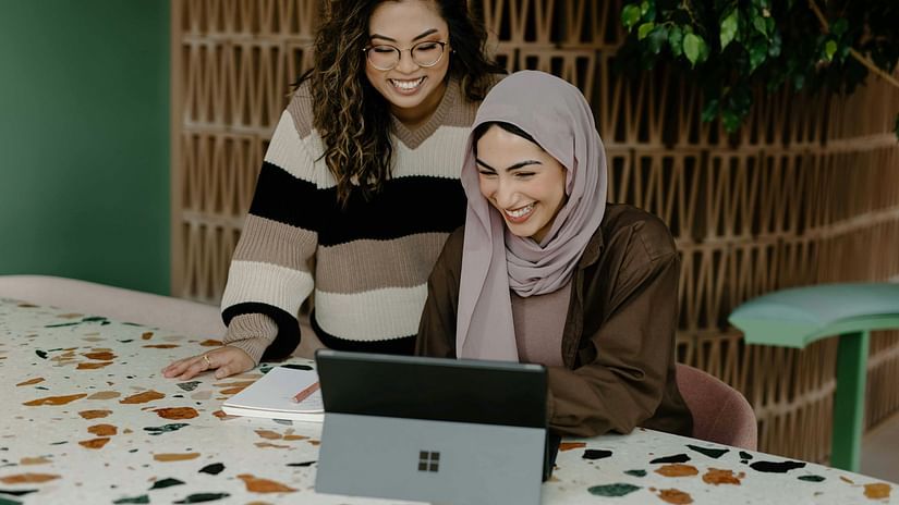 Two women working in front of a laptop while smiling inside a work place.