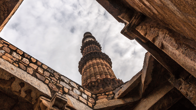 A captivating low-angle view of Qutb Minar tower in Delhi, India, surrounded by rustic stone walls under a cloudy sky.