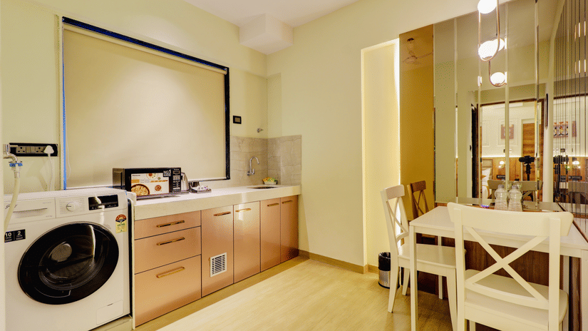 The interior at  Home2 India, with washing machine, cabinets, countertop, and sink, with a small dining table and chairs placed near a glass partition.