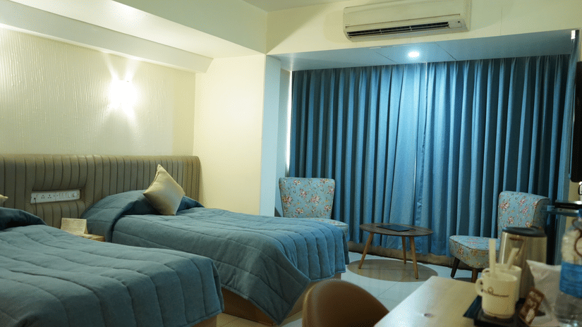 A room with twin beds, spreads, pillows, chairs, and a large window covered with curtains.
