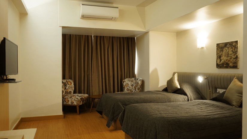 A room with twin beds, spreads, pillows, a television, and a large window covered with curtain.