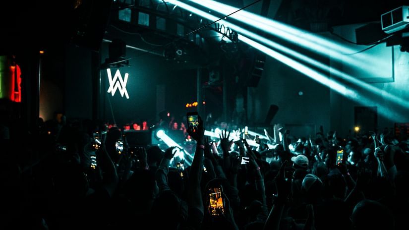  A packed nightclub with a large crowd facing a stage with a prominent AW logo. Multiple spotlights illuminate the scene, and the audience is holding up their phones, capturing the moment.