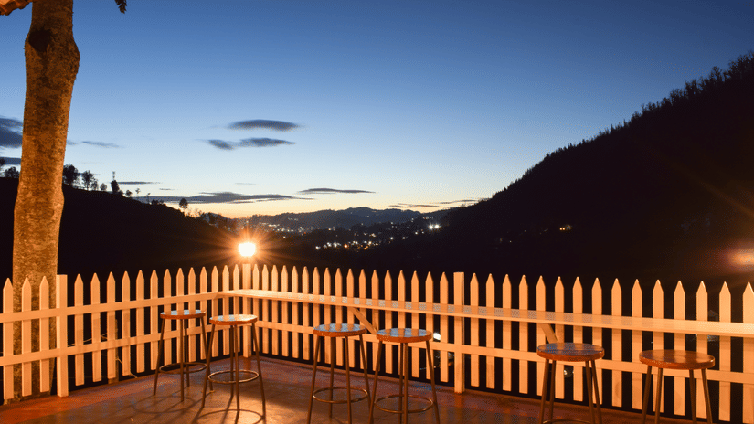 An outdoor patio with a white picket fence at dusk or dawn, featuring tables and chairs, and a distant view of rolling hills under a gradient sky.