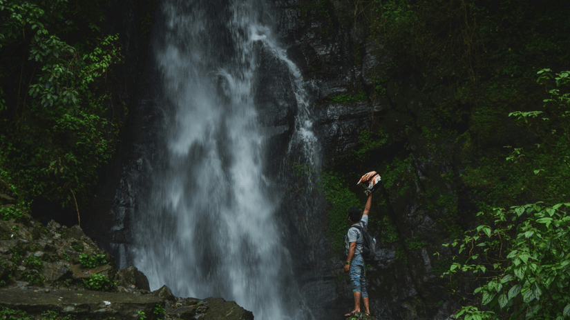 A person with a rucksack stands before a tall, misty waterfall cascading down a rocky cliff in a lush green forest.