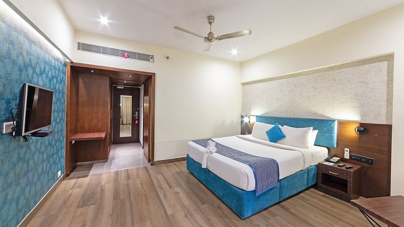 A corner spacious and well-lit hotel room with a bed, a flat-screen TV, and a seating area. The room features a wooden floor and blue accents on the walls.