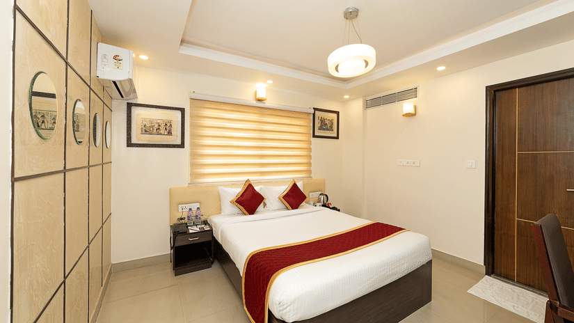 The Deluxe Room of Icon Regency by Bhagini, Whitefield showcasing a double bed with crisp linens, wardrobe, an air conditioning, ceiling lights, closed wooden door, and tiled flooring.