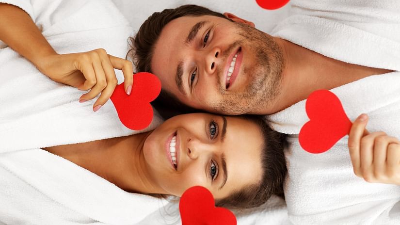 A smiling couple wearing white bathrobes holding red paper hearts on each hand, laying with their heads aligning adjacently opposite to each other's.