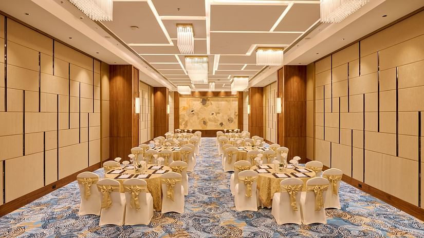 A spacious function room is set up for an event, with rows of round tables draped in white cloths and adorned with gold sashes on the chairs, under modern rectangular lighting. - Iris Sarovar Portico, Sector 85, Gurugram
